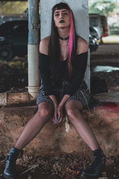 goth fashion style classic; girl  with pink streak in hair, heavy eye makeup, black top, black plaid skirt and fishnet tights