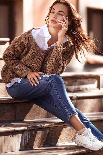 sophisticated woman in white blouse with tan knit sweater over with fitted jeans and white sneakers, exuding timeless style outfit idea