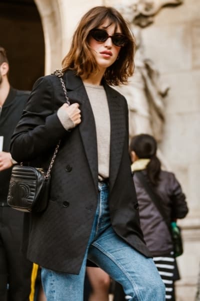  jeanne damas à paris vêtue d'un pull, d'un blazer structuré, de lunettes de soleil, d'un sac chanel et d'un jean à jambe droite