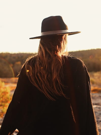 woman with large brim fedora and loose soft-edged coat walking at sunset to exude flamboyant natural aesthetic 