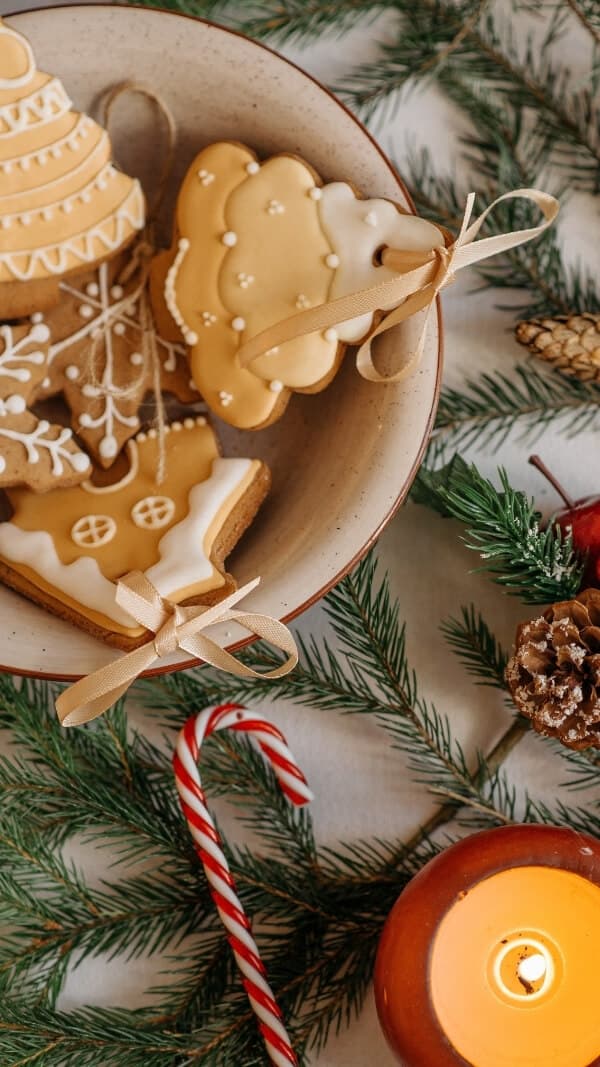 flatlay image of evergreen sprigs, candy canes, a candle, and a bowl of gingerbread cookies- soft, warm colors, holiday christmas phone background 