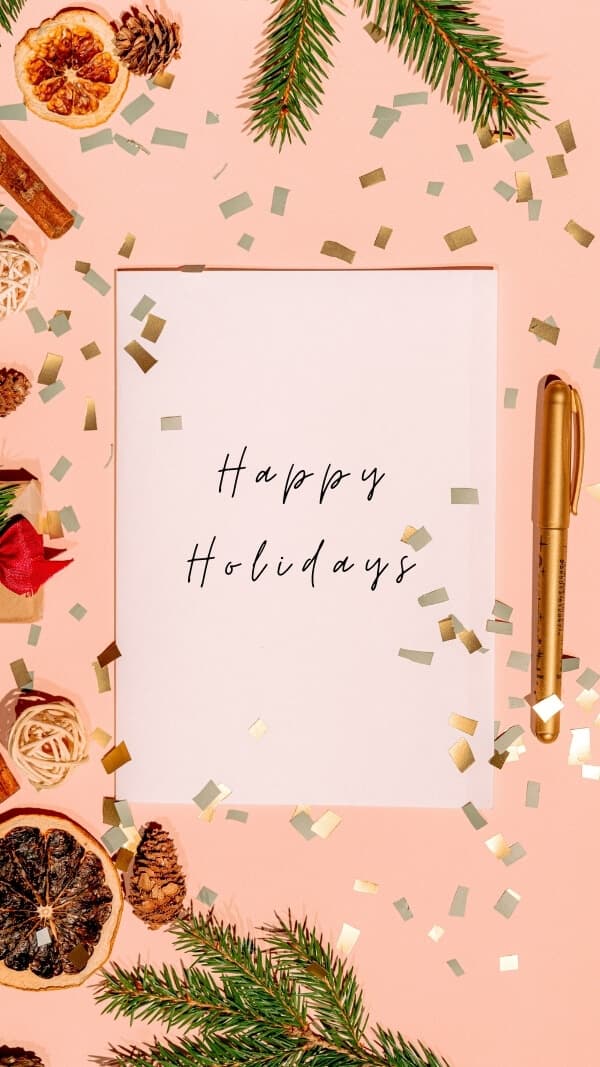 peach background with holiday flat lay elements confetti, dried oranges, evergreen sprigs, and a piece of paper in center with "happy holidays" on it