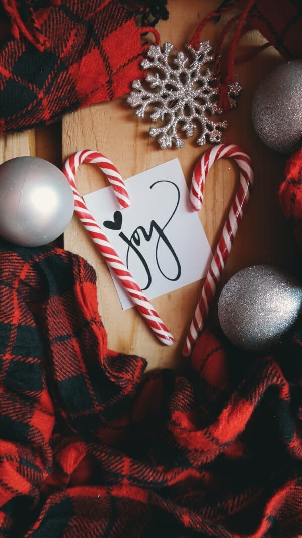 holiday flatlay with red plaid fabric, cand canes, and ornaments with a post it in center with the words "joy" on it, christmas holiday wallpaper background iphone