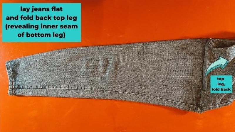 lay jeans flat and fold back top leg (revealing inner seam of bottom leg)- arrow showing top leg being folded up towards waistband 