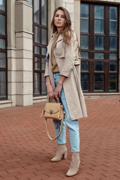 woman wearing classic trench with sweater and jeans and heeled tan boots and bag